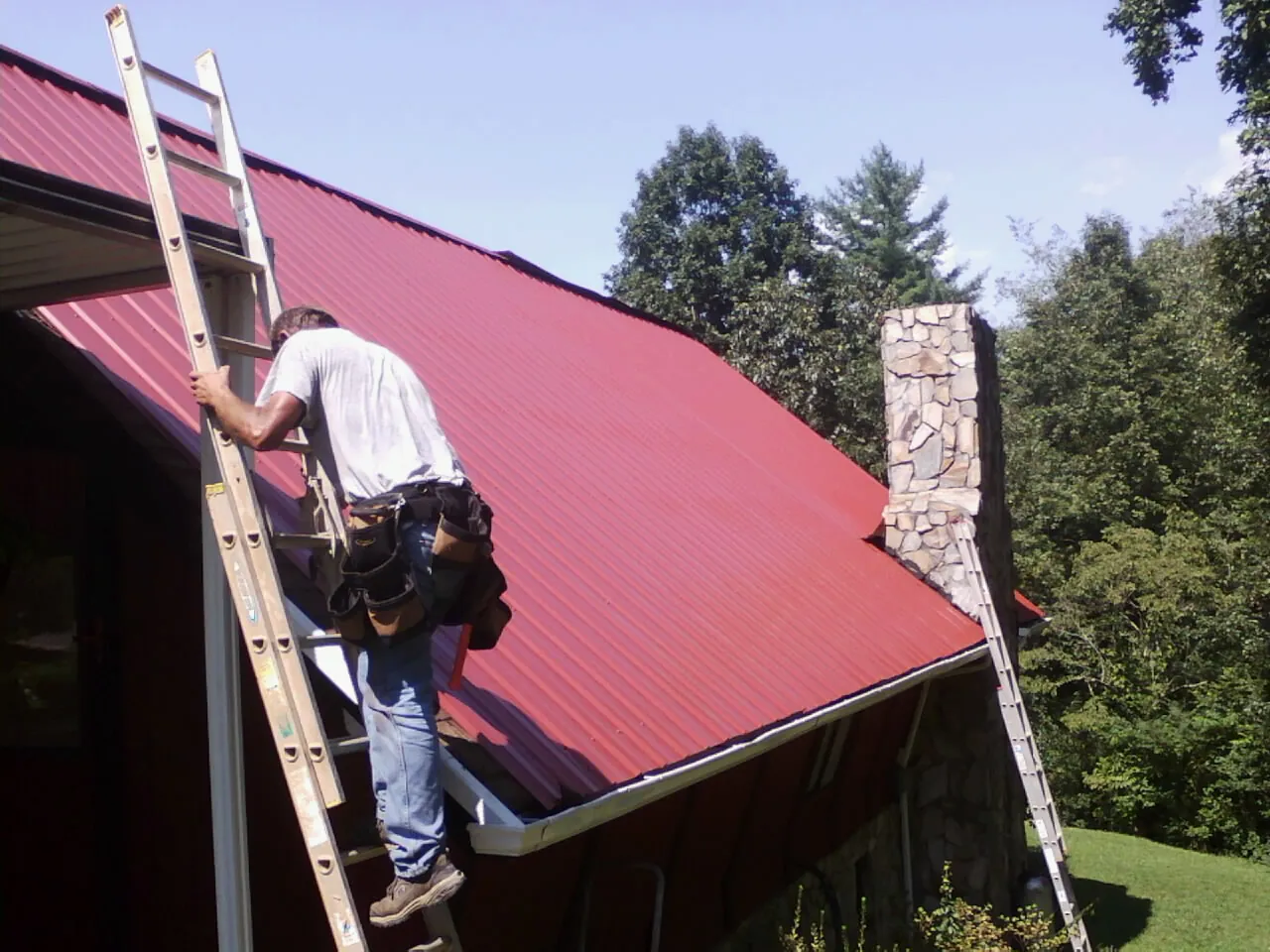Roofer on a ladder beside a red metal roof and stone chimney — Soffit & Fascia Repair by Roofing Brothers in Adams Run
